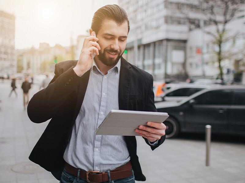 A man using a tablet and talking on a phone as he walks in the city